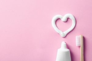A tube of toothpaste and a toothbrush under a heart drawn with toothpaste on a pink background 